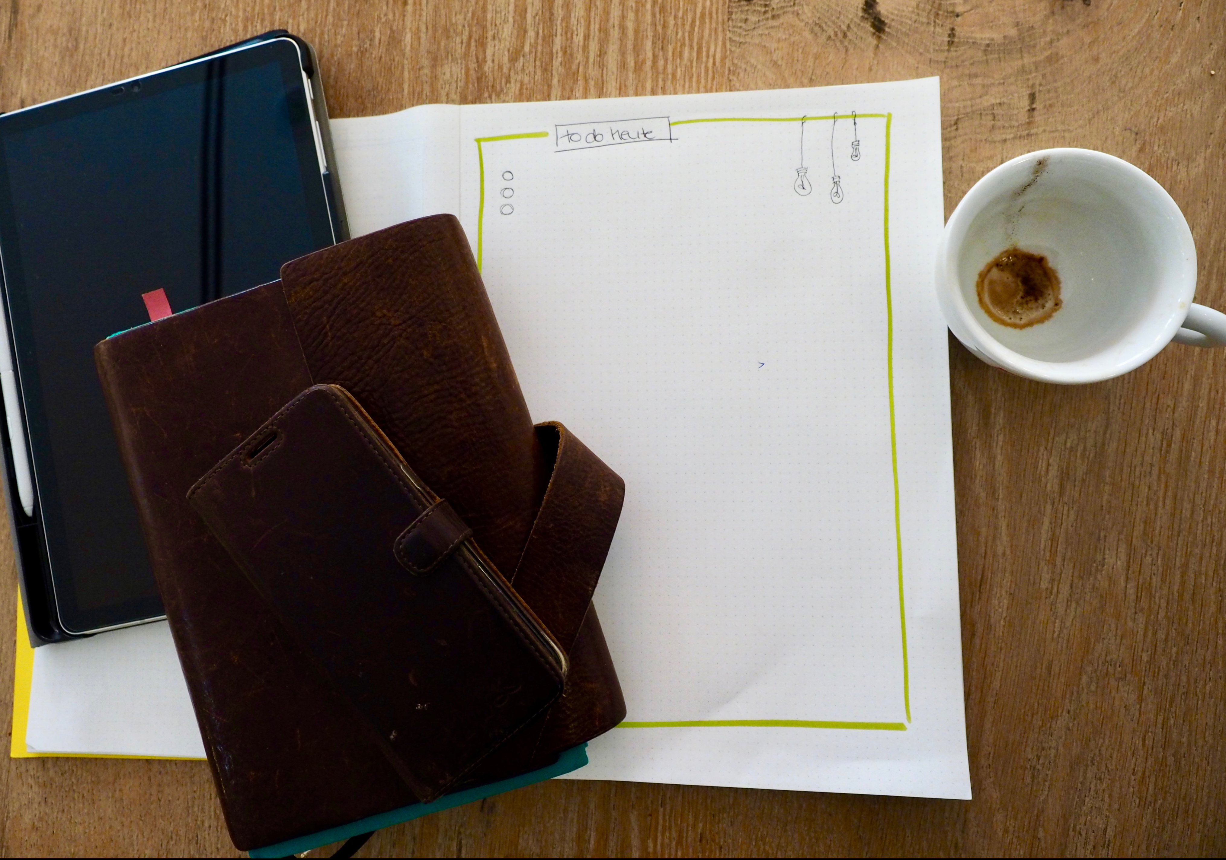 Frau Bevers Arbeitsmaterialien – ein Block, Tablet, Smartphone und Kalender – liegen neben einer Kaffetasse auf einem Holztisch